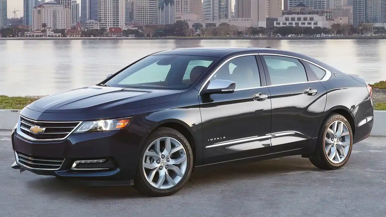 A black Chevrolet Impala sedan parked near a waterfront with a city skyline in the background, showcasing its smooth design and spacious interior.