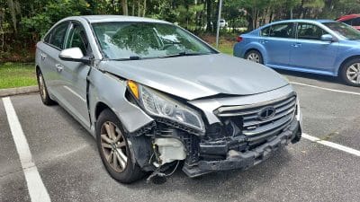 A silver sedan with significant front-end damage, including a crumpled hood, broken headlight, and damaged bumper, parked in a parking lot.