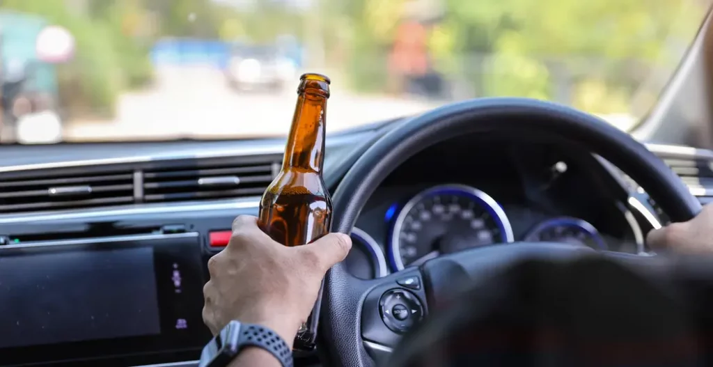 Driver irresponsibly holding a beer bottle while operating a car, exemplifying the dangerous behavior targeted by NHTSA's proposed in-car detection systems.