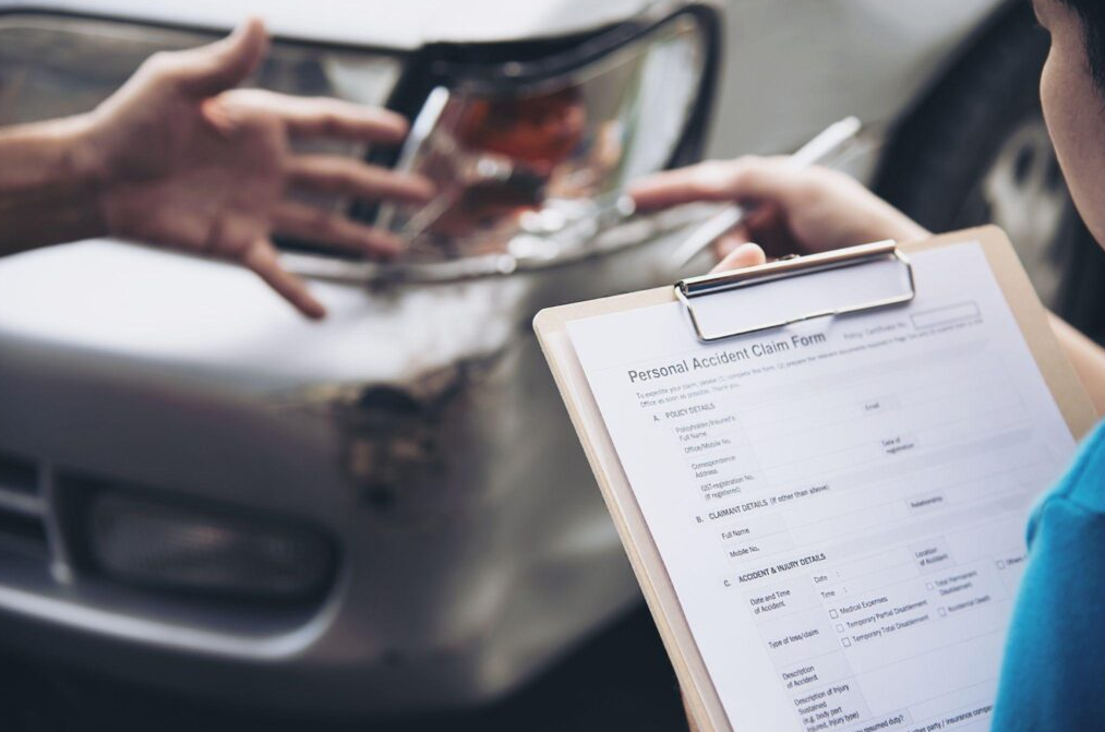 Person filling out accident claim form next to damaged car – Diminished Value of Georgia, Car Appraisals for Insurance Claims Insurance adjuster holding a personal accident claim form next to a damaged vehicle for a diminished value claim