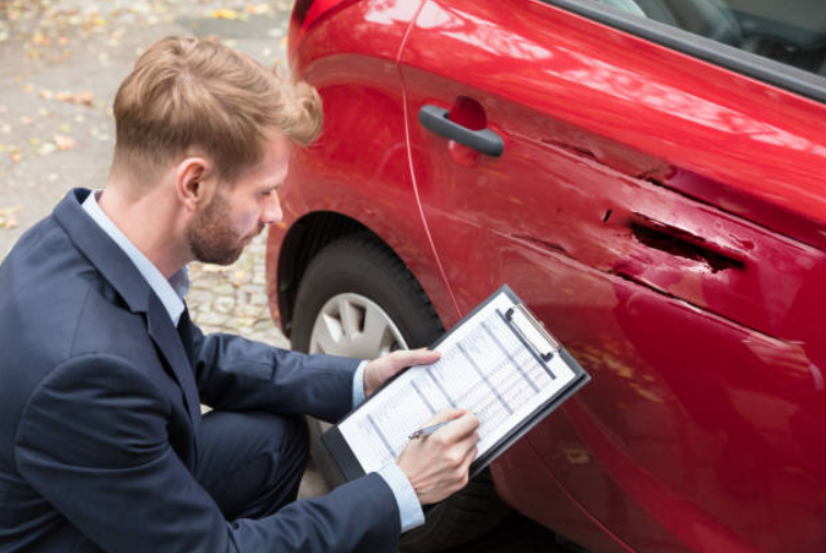 insurance-adjuster-inspecting-car-damage-georgia-diminished-value-claim – Diminished Value of Georgia, Car Appraisals for Insurance Claims Man in a suit inspects a scratched red car door and records details on a clipboard outside.