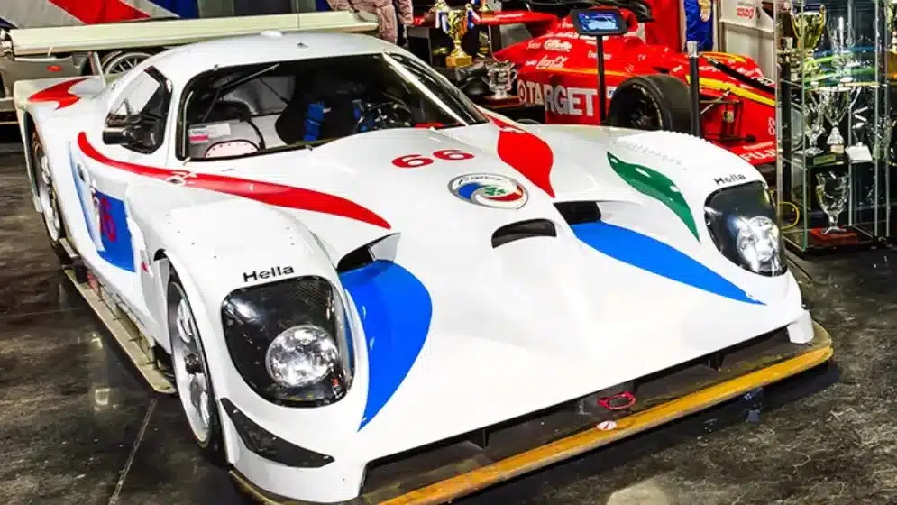 A Panoz race car, emblazoned with the number 66 and featuring a striking white body with red, blue, and green accents, is showcased in a museum-like setting. The car is surrounded by racing memorabilia, including trophies and racing suits, highlighting its storied history in motorsport. Behind it, another race car and more trophies are displayed, emphasizing the legacy of Panoz in competitive racing.