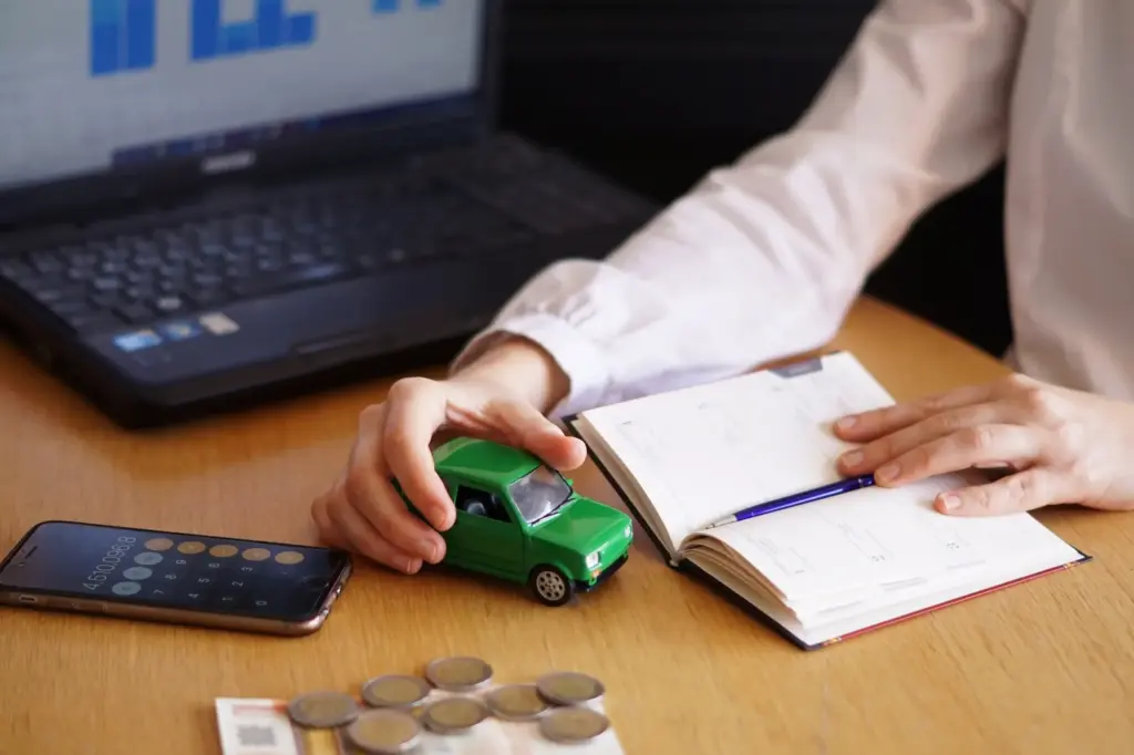 A person calculating car-related expenses, holding a small green toy car, with coins, a smartphone calculator, and a notebook on the desk, and a laptop in the background.